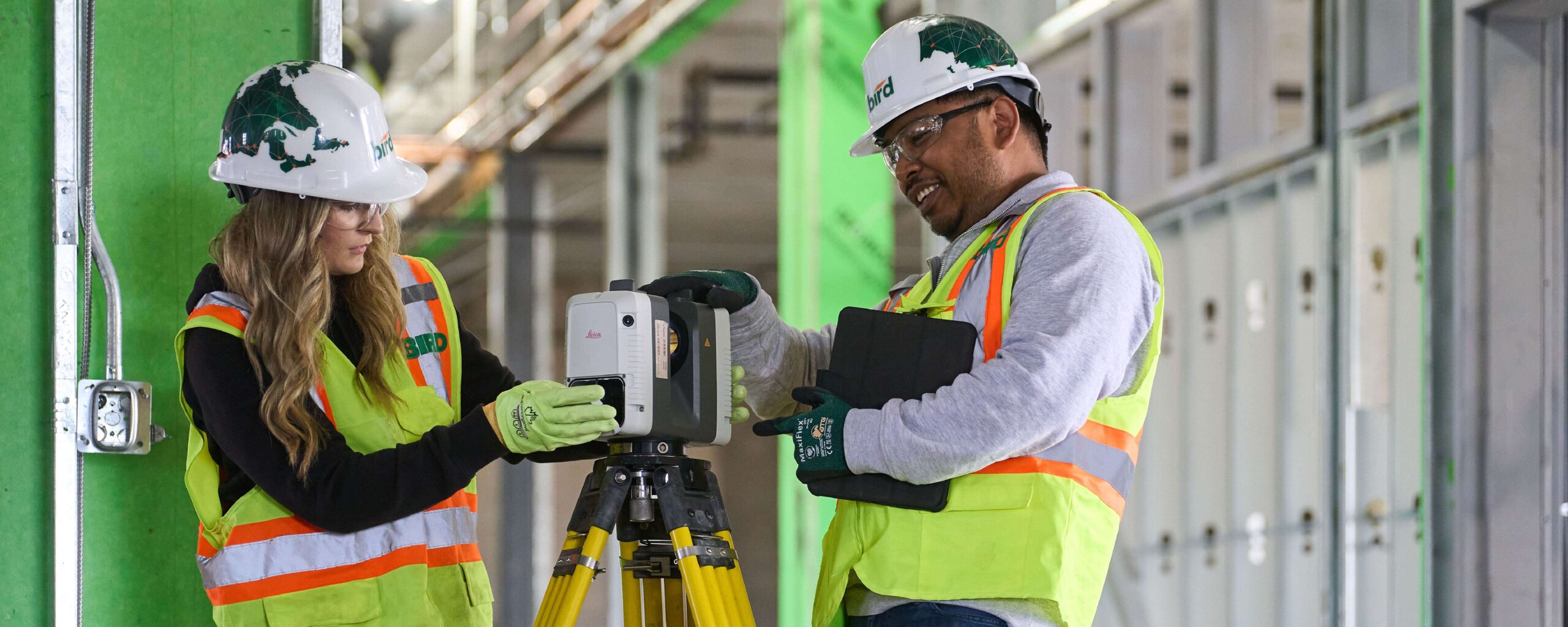 Website Banners_1500x600_Careers 1 Two construction workers wearing hard hats and safety vests use a surveying instrument on a tripod inside a building under construction. One holds the device, while the other looks at a tablet.