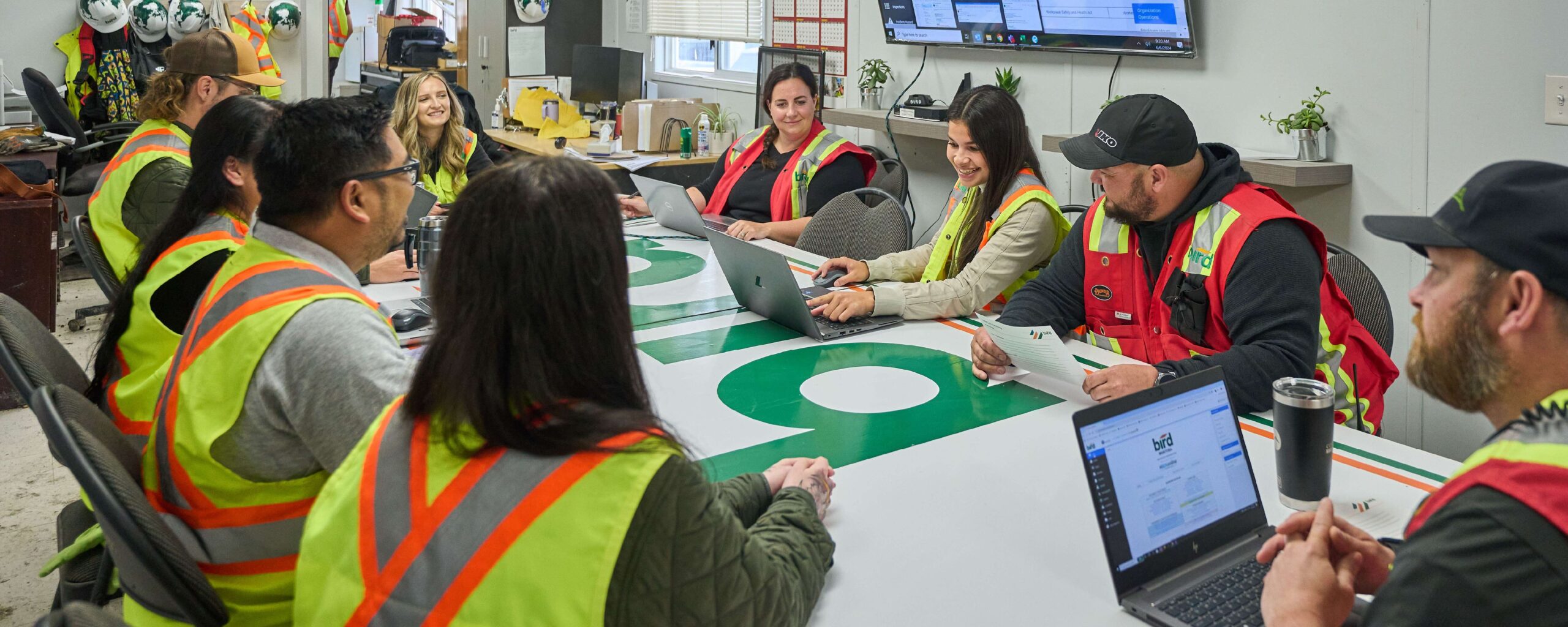 Website Banners_1500x600_Culture-Safety 1 A group of construction workers wearing safety vests sit around a table in a meeting room, discussing and using laptops and papers, with safety gear and monitors visible in the background.