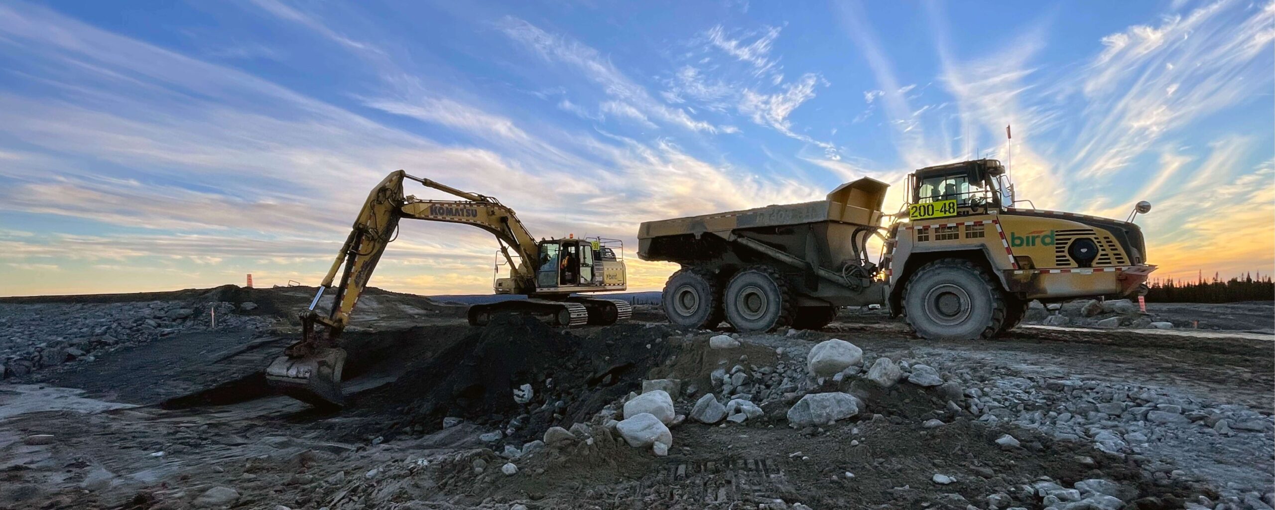 Website Banners_1500x600_Industrial Excavator loading soil into a large dump truck at a construction site during sunset, with a dramatic sky and scattered clouds in the background. Rocks and dirt cover the ground.