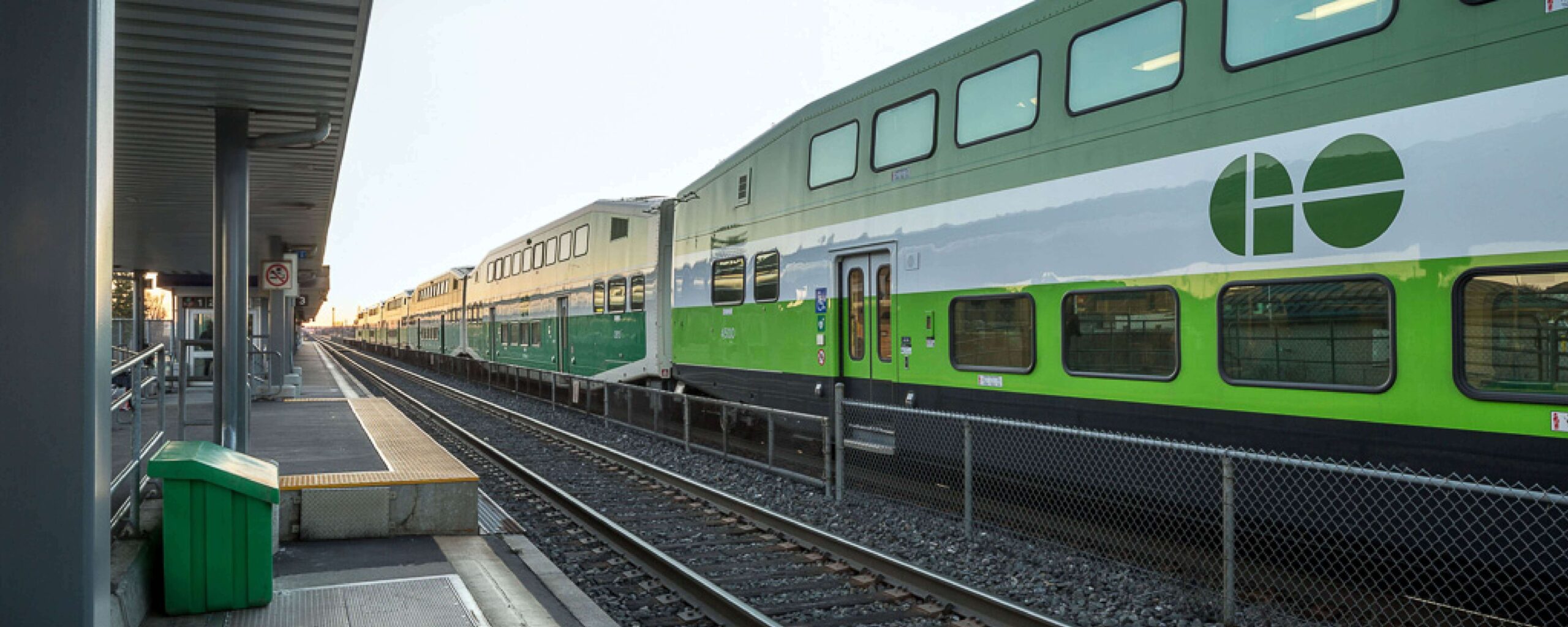 Website Banners_1500x600_Infrastructure A green and white double-decker GO Transit train is stopped at an outdoor station platform, with empty tracks and a fence running alongside the train.
