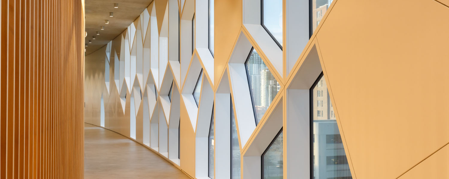 Website-Banners_1500x600_Buildings A modern hallway with a curved wall of large, geometric, diamond-shaped windows letting in natural light; wood paneling lines the opposite wall and ceiling. City buildings are visible through the windows.
