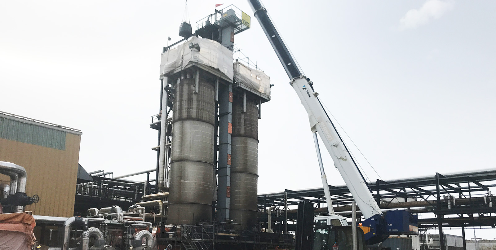 Un chantier de construction avec une grande structure industrielle comprenant deux grands réservoirs cylindriques, une grue soulevant des matériaux et divers tuyaux. La zone est délimitée par un sol en terre battue et des barrières de sécurité rouges.