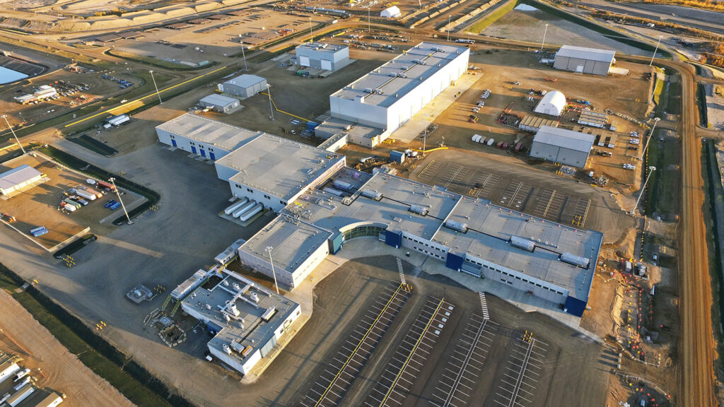 Vue aérienne d'une grande installation industrielle comprenant plusieurs bâtiments reliés entre eux, des parkings et les routes environnantes. La zone comprend des véhicules garés, des terrains découverts et des entrepôts à proximité, avec un terrain dégagé et généralement plat autour du site.