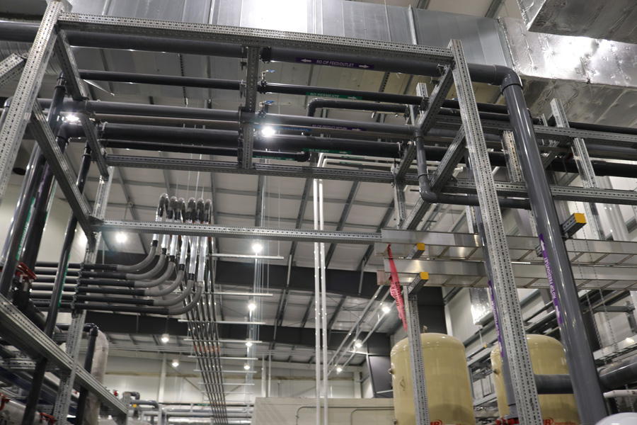 Industrial interior with metal pipes and ducts running along the ceiling and walls. The space is brightly lit, with structural beams and support frames visible. Tanks and machinery are partially seen in the background.