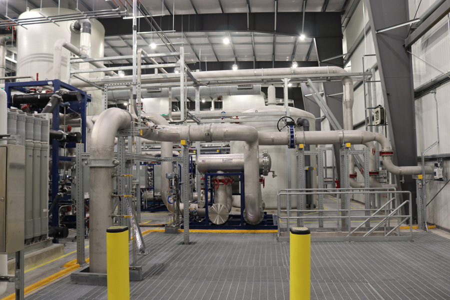 Industrial facility interior with large metal pipes, tanks, and machinery. The space has a high ceiling, overhead lighting, safety railings, and yellow bollards, indicating a controlled and organized environment.