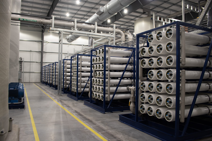 Rows of large cylindrical tanks and piping in an industrial facility, likely part of a water treatment or desalination plant, with clean concrete floors and a high ceiling with visible ductwork.