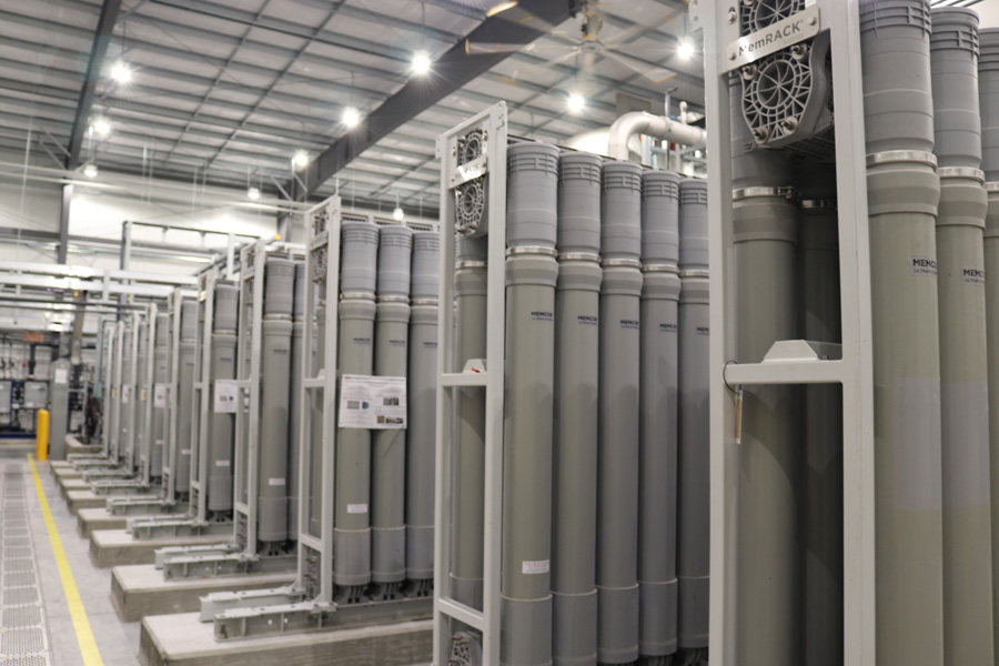 Rows of large, vertical cylindrical industrial filters or membranes are arranged in a spacious, well-lit factory or facility with a high ceiling and metal framework.