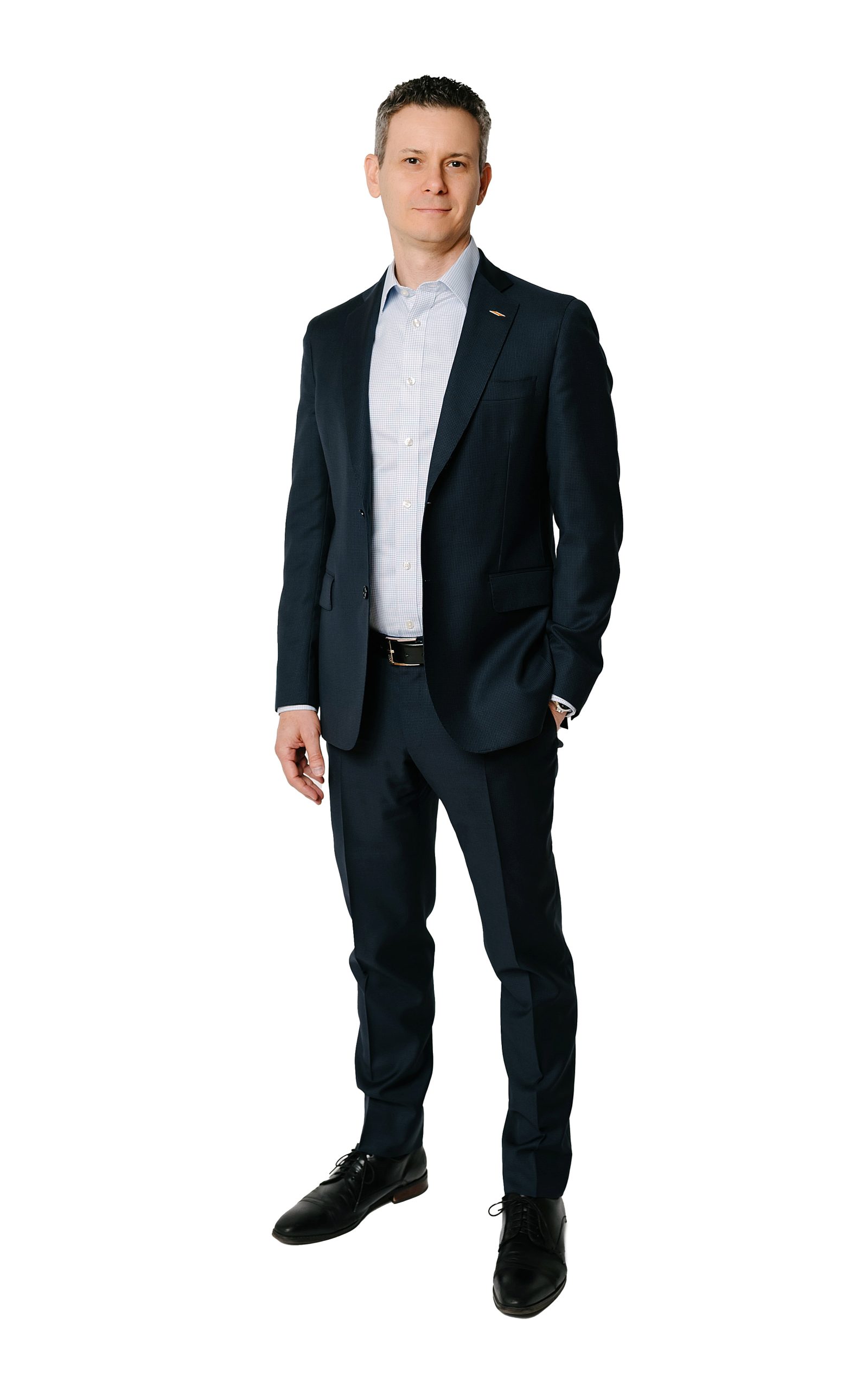 A man wearing a dark suit, light dress shirt, and dress shoes stands confidently against a plain white background, looking at the camera with a slight smile.