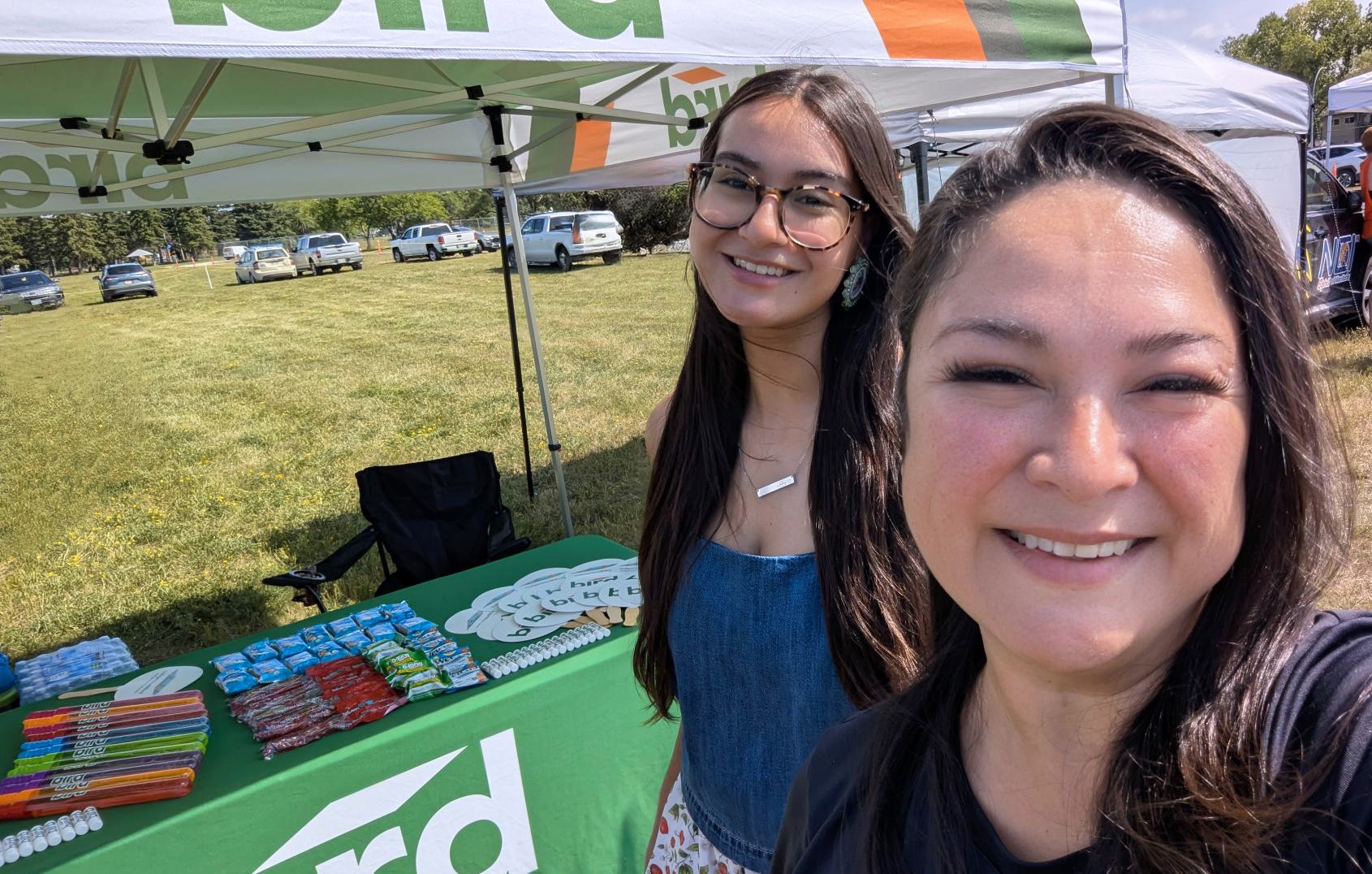 Two women smile in front of a green booth table covered with colorful snacks and branded items at an outdoor event, with cars and trees in the background.