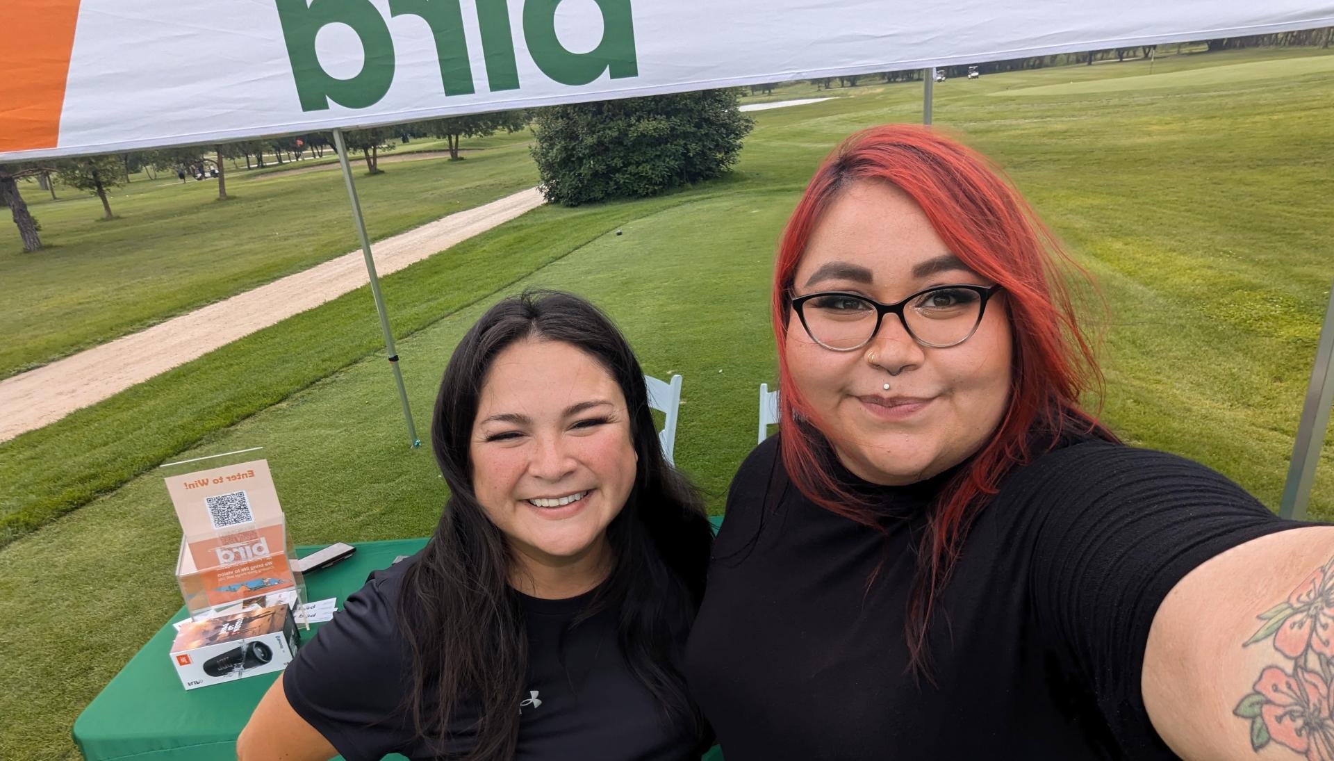 Two women smiling and posing for a selfie under a canopy at an outdoor event, with green grass, trees, and a table with Bird-branded materials in the background.