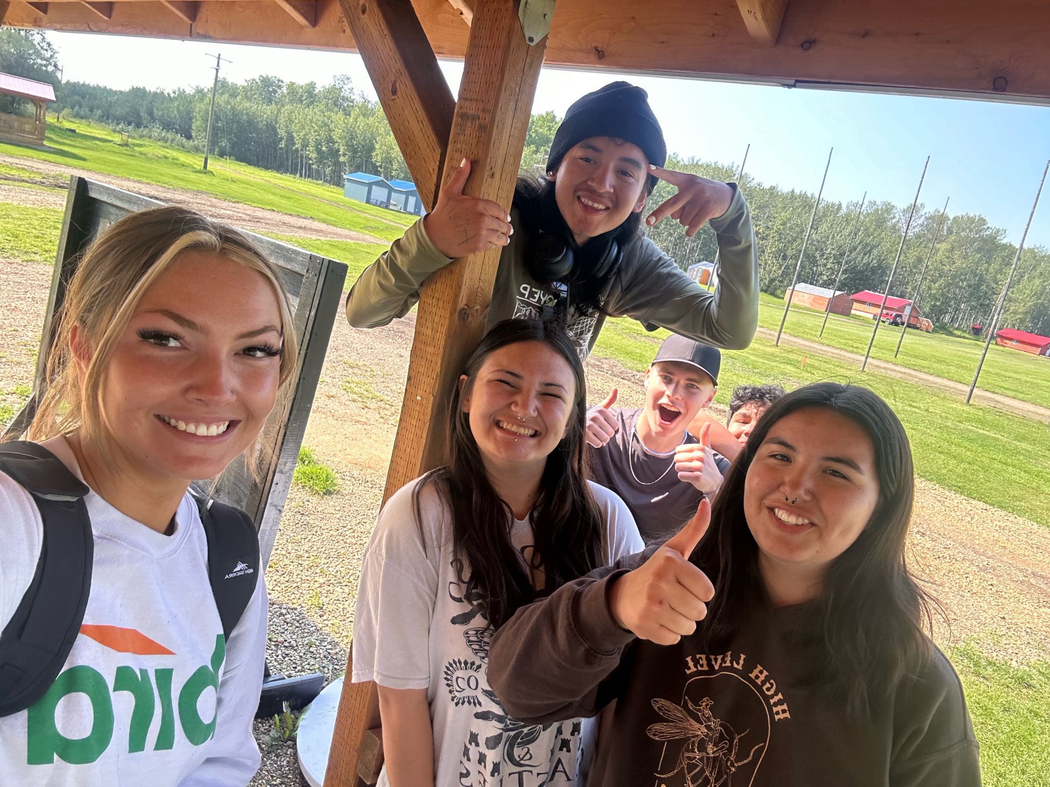 A group of five smiling teenagers pose for a selfie outdoors under a wooden shelter; one person stands above, others stand together, and everyone looks happy, giving thumbs up signs. Green grass and trees are visible in the background.