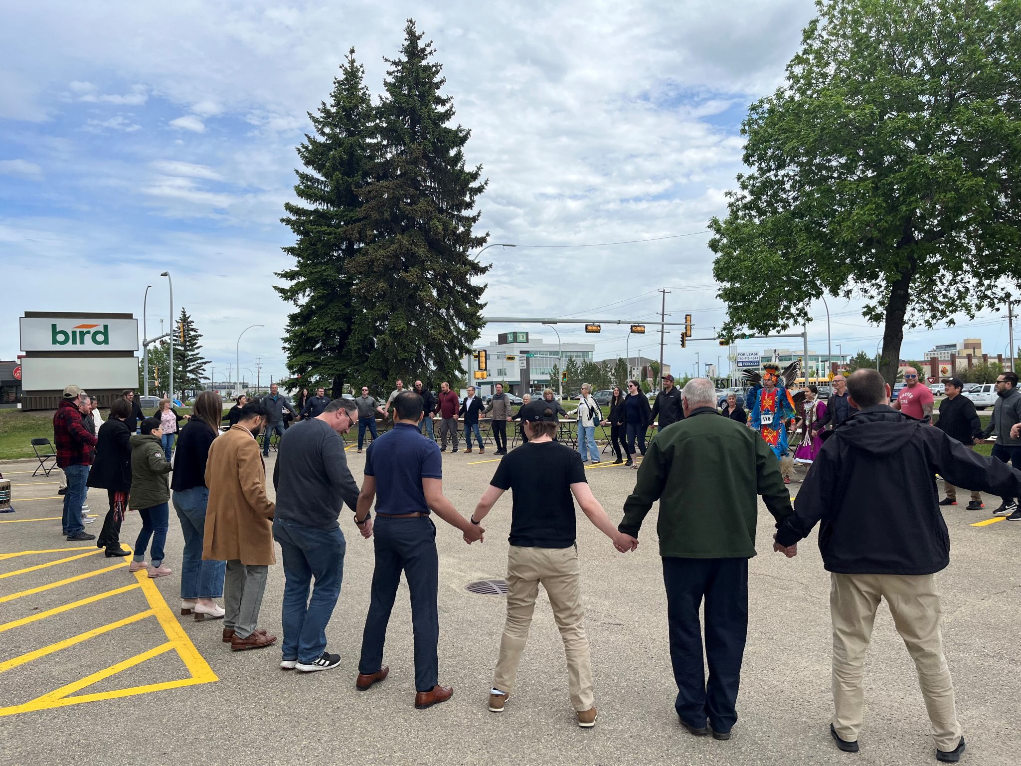 A large group of people stand in a circle holding hands in a parking lot, participating in a communal outdoor event under a cloudy sky, with trees and buildings in the background.