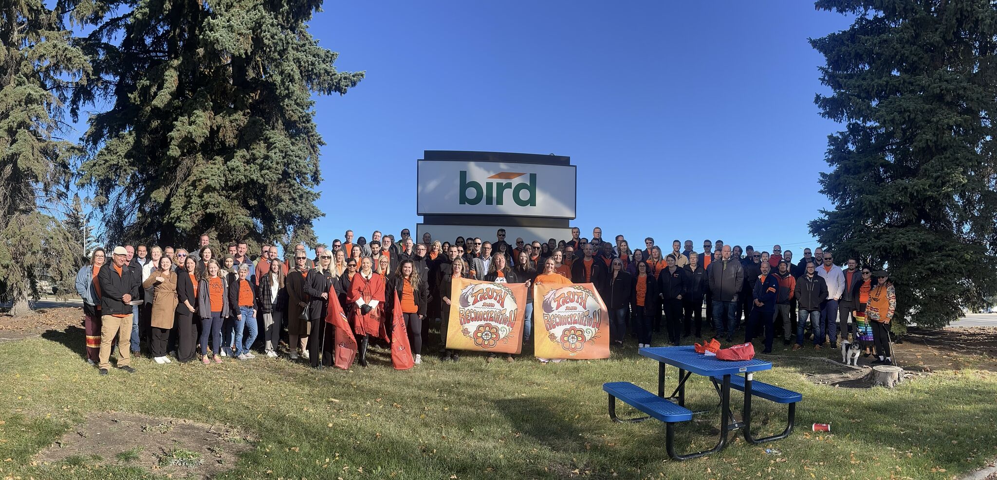 A large group of people pose together outdoors in front of a bird sign, standing on grass with tall evergreen trees behind them. Two banners with fall-themed designs are displayed at the front. A blue picnic table is visible.