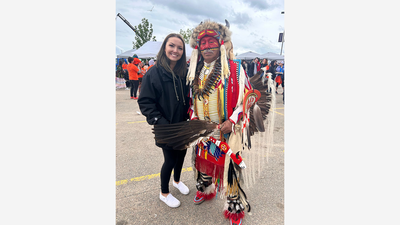 A woman in casual clothing stands next to a man wearing traditional Indigenous regalia with feathered accessories, beadwork, and a headdress at an outdoor event with tents in the background.