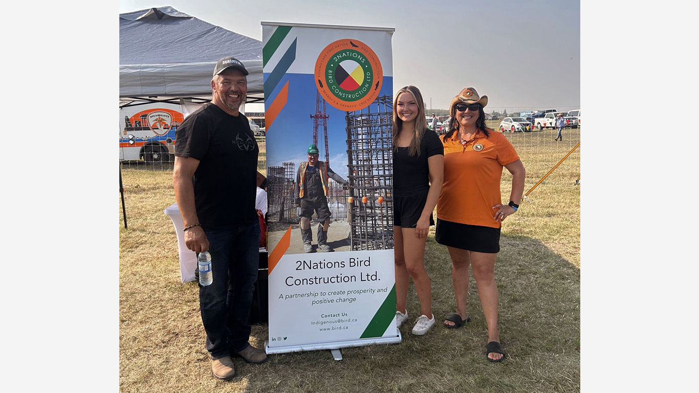 Three people smile and stand next to a 2 Nations Bird Construction Ltd. banner at an outdoor event. The banner features a construction worker and the company logo. The group is on a grassy field with tents and people in the background.