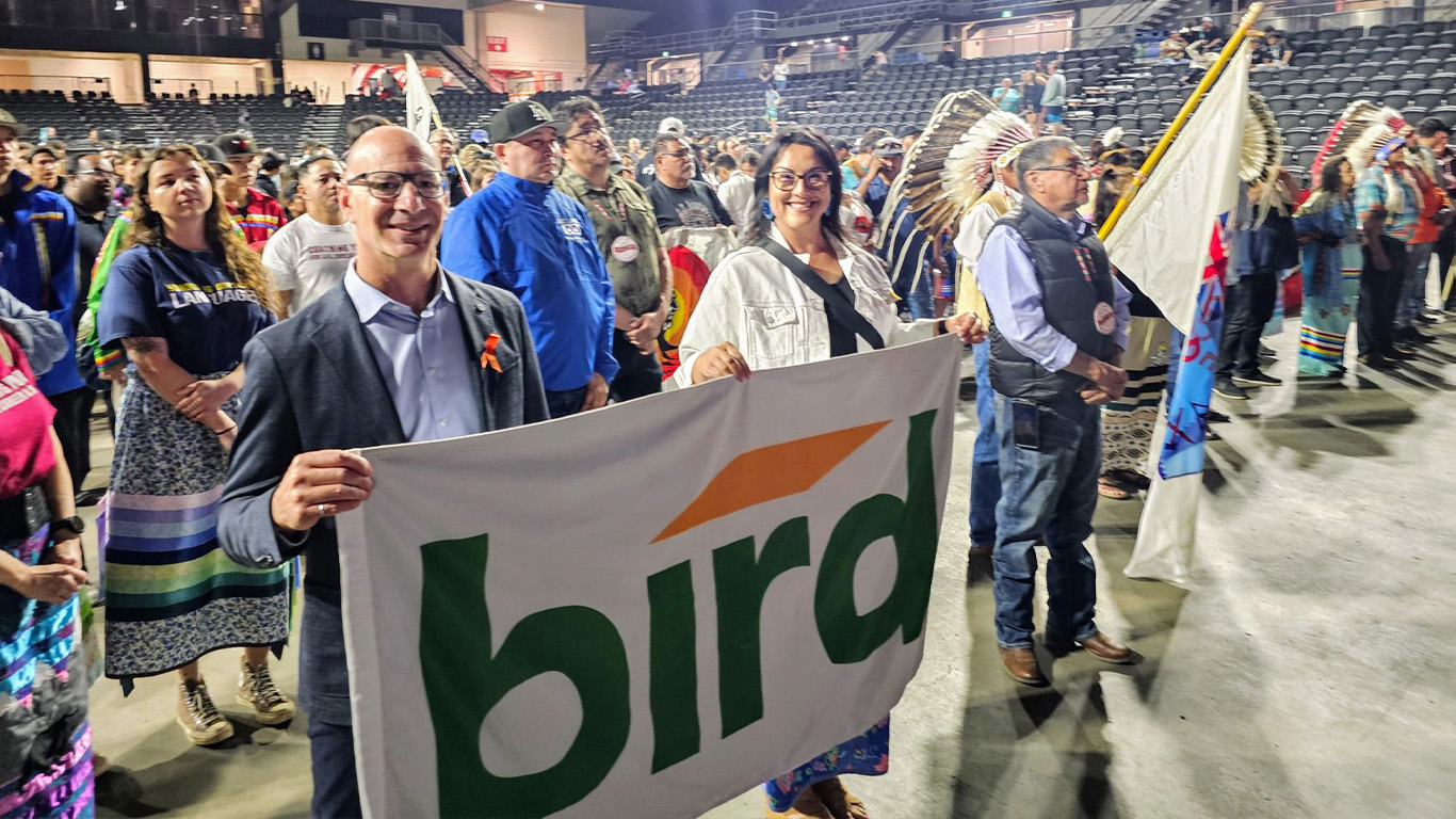 A group of people stand in an indoor arena, some holding flags and wearing traditional attire. Two people in front smile and hold a bird banner. The seats behind them are mostly empty.