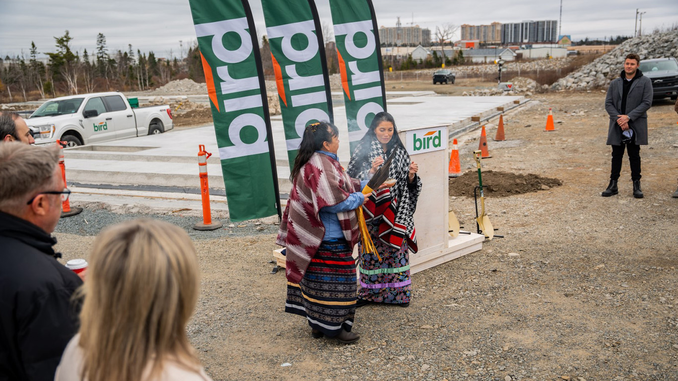 Two women in traditional attire perform a ceremony at a construction site, surrounded by people. Three green Bird flags, shovels, and a Bird truck are visible in the background.