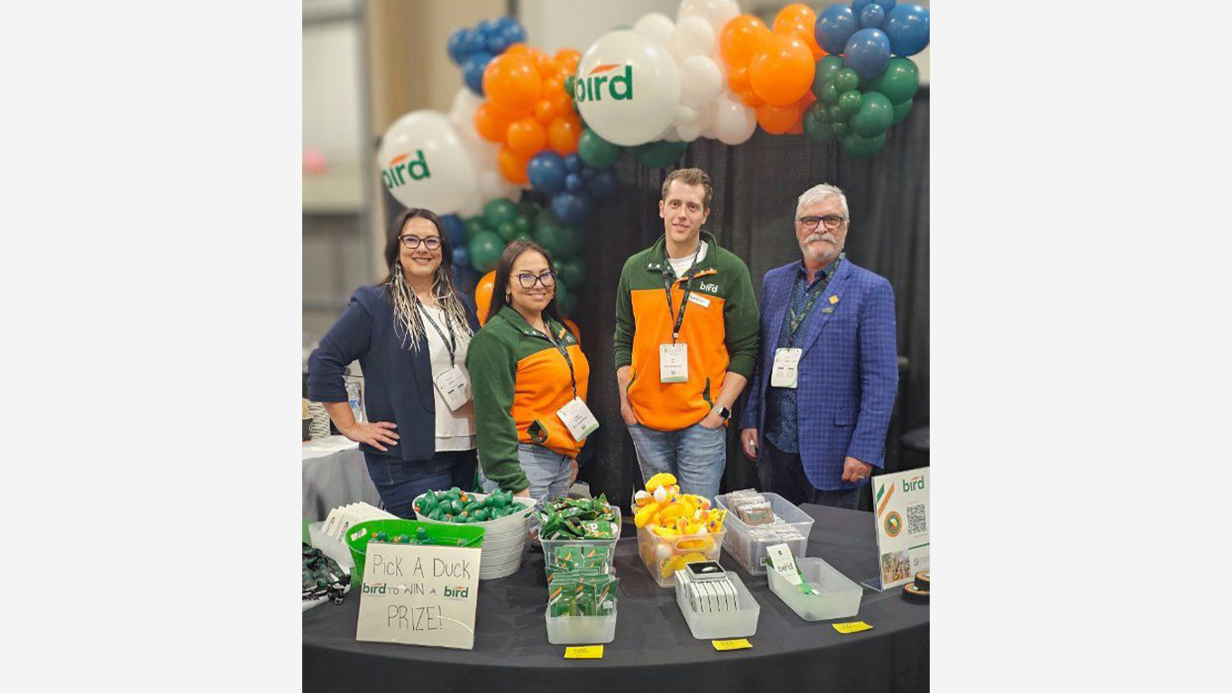 Four people stand behind a table with Bird branding at a booth decorated with colorful balloons. The table displays toy ducks, prizes, and a sign reading “Pick a Duck bird to WIN a bird PRIZE!”.