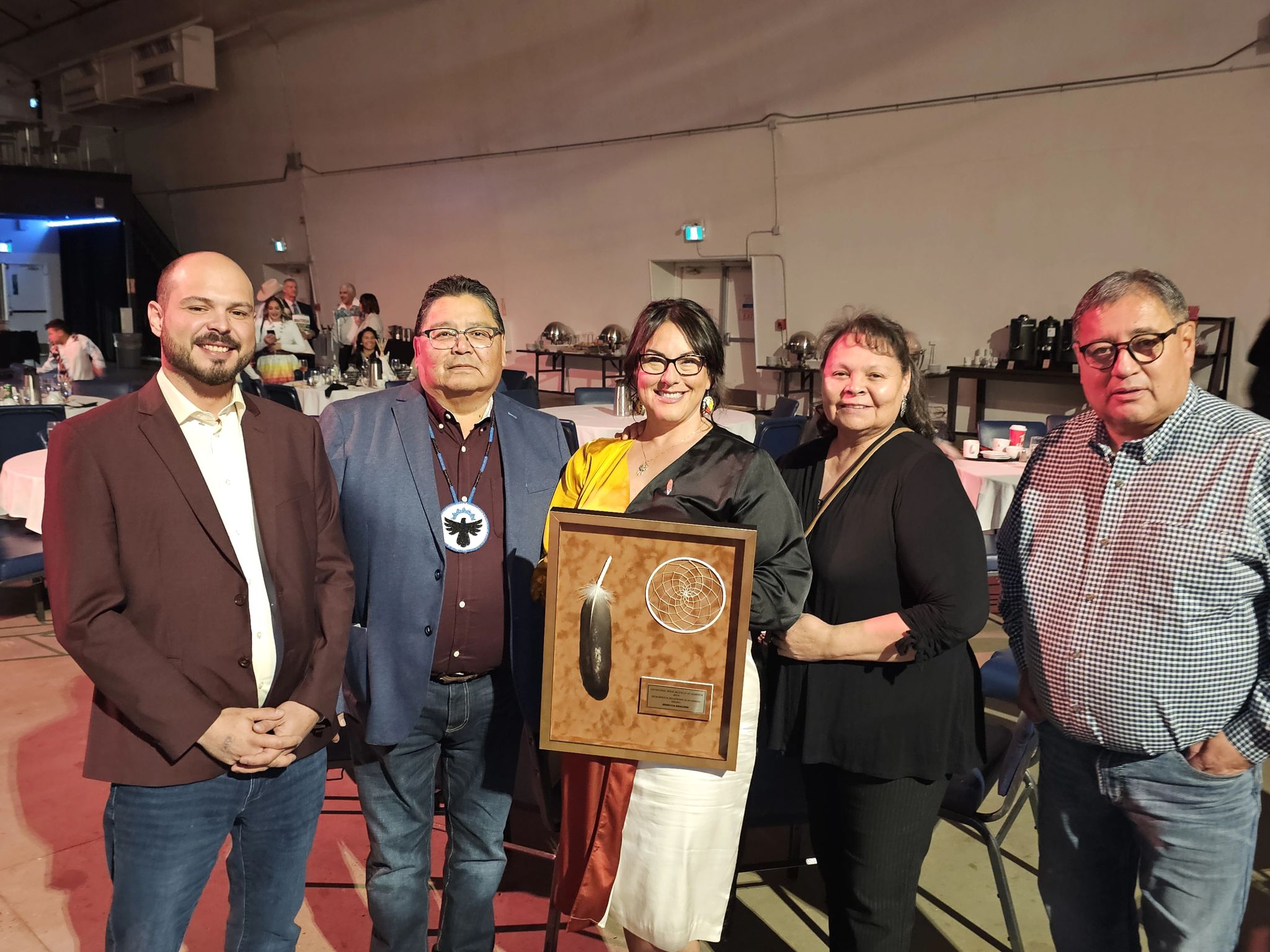 Five adults stand together indoors at an event. The woman in the center holds a framed award featuring a feather and two circular designs. Tables and people are visible in the background. Everyone is smiling.