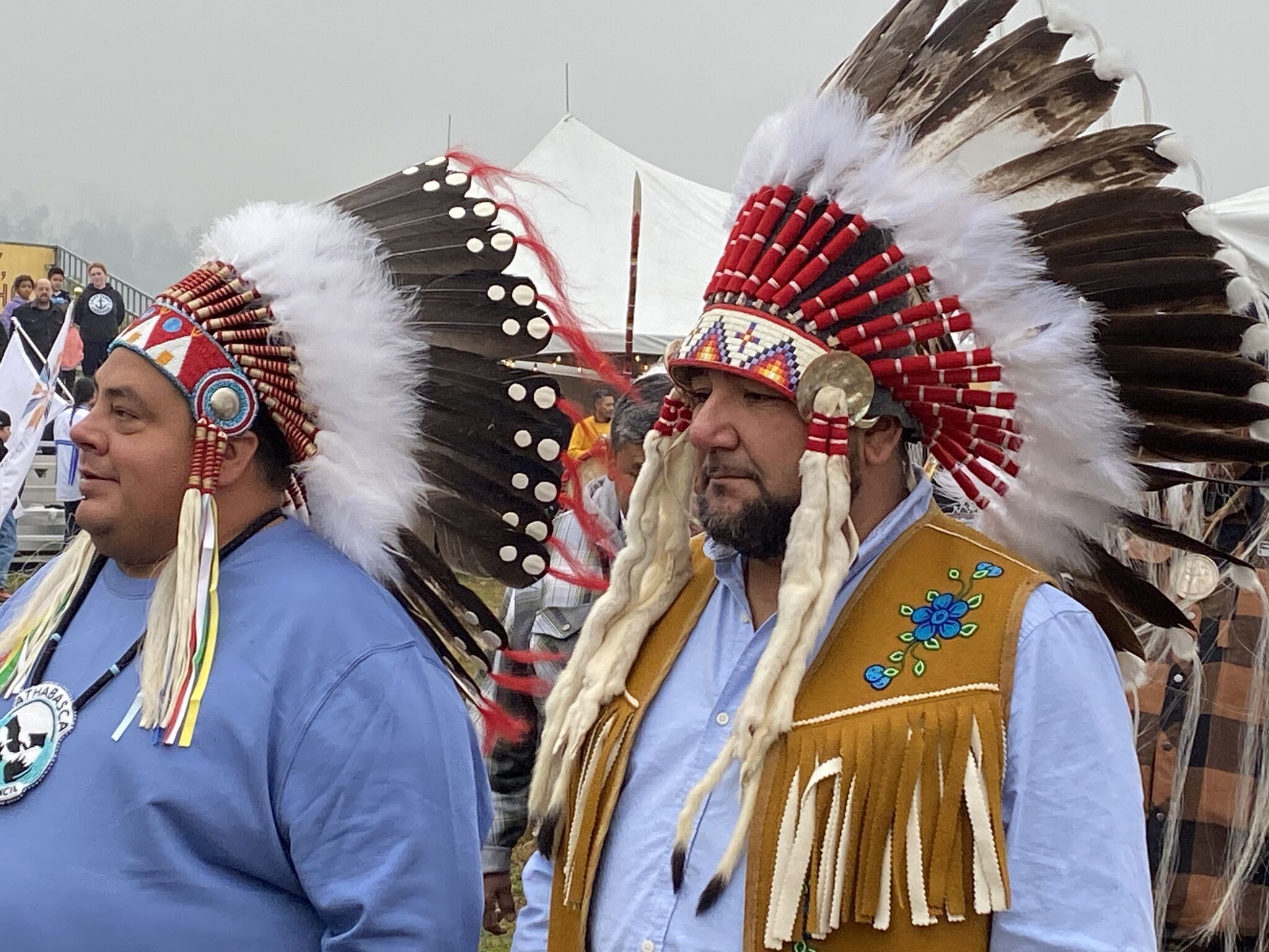Two men wear traditional Indigenous regalia, including feathered headdresses and beaded accessories, standing outdoors with tents and a crowd in the background on a foggy day.