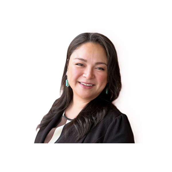 A woman with long dark hair smiles at the camera. She is wearing turquoise earrings, a black blazer, and a patterned top, posed against a plain white background.
