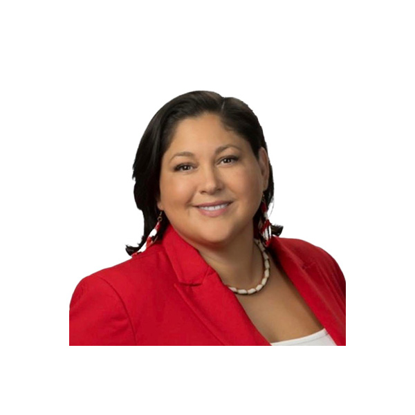 A woman with dark hair wearing a red blazer, white top, beaded earrings, and a white necklace, smiling at the camera with a plain white background.