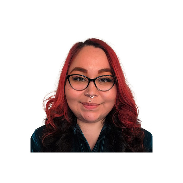 A person with long, wavy red hair, wearing black cat-eye glasses and a dark top, is smiling at the camera. They have a septum piercing and groomed eyebrows, with a white background behind them.
