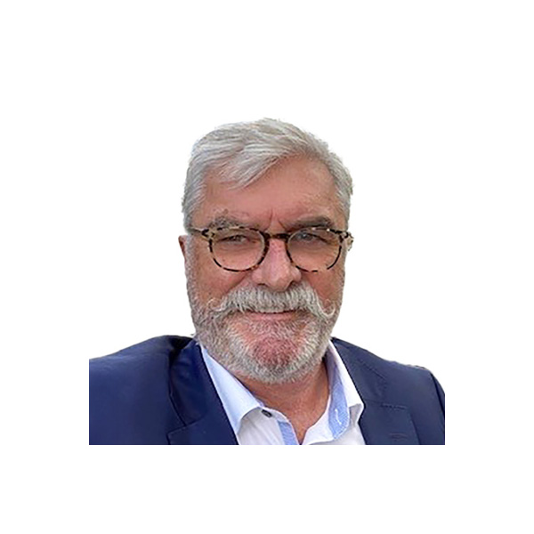 A man with gray hair, a mustache, and beard, wearing glasses and a dark blazer over a white shirt, is smiling at the camera against a plain white background.