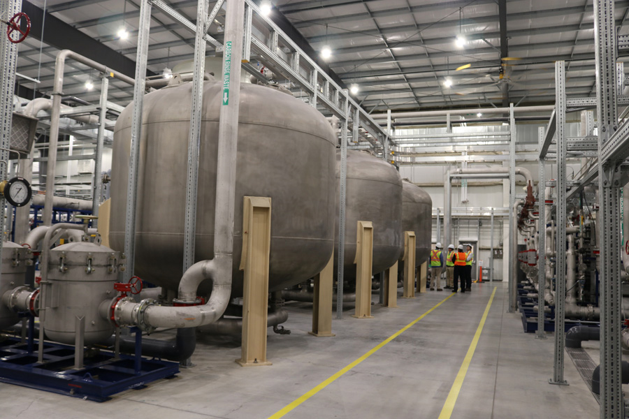 Large industrial water treatment tanks and pipes inside a spacious facility, with a few workers wearing safety vests and helmets inspecting the equipment. The area is well-lit with a metal structural ceiling.