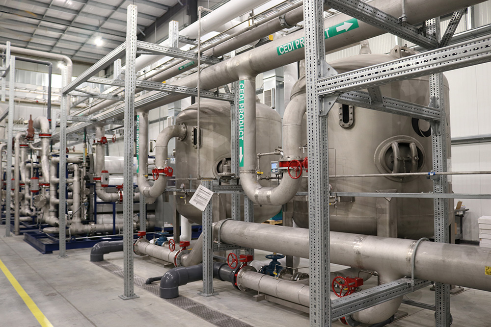 Large industrial pipes and tanks inside a modern facility, surrounded by metal supports and valves, with labels and safety signs visible on the equipment. The environment is clean and well-lit.