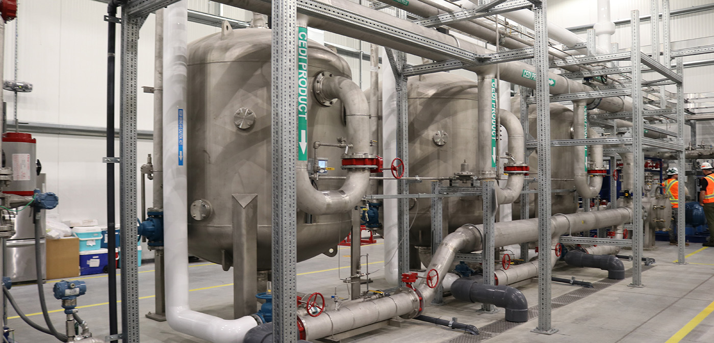 Industrial facility interior with multiple large metal pipes, cylindrical tanks, and machinery arranged in rows. Overhead pipes run horizontally, and various gauges and control panels are visible in the background.
