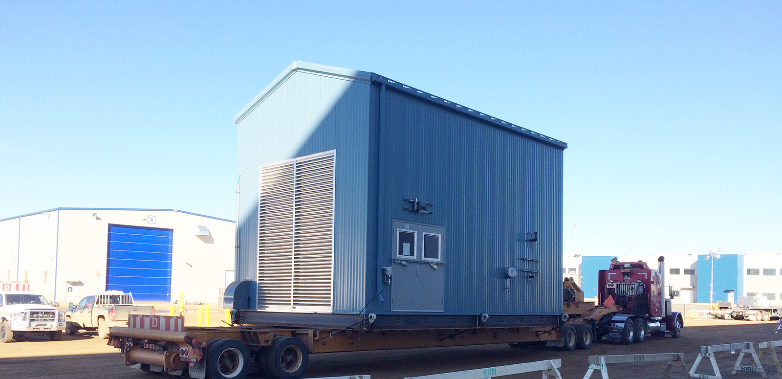 A large, silver industrial building sits elevated on wooden blocks and metal supports in a snowy lot, surrounded by orange cones and equipment under a clear blue sky.