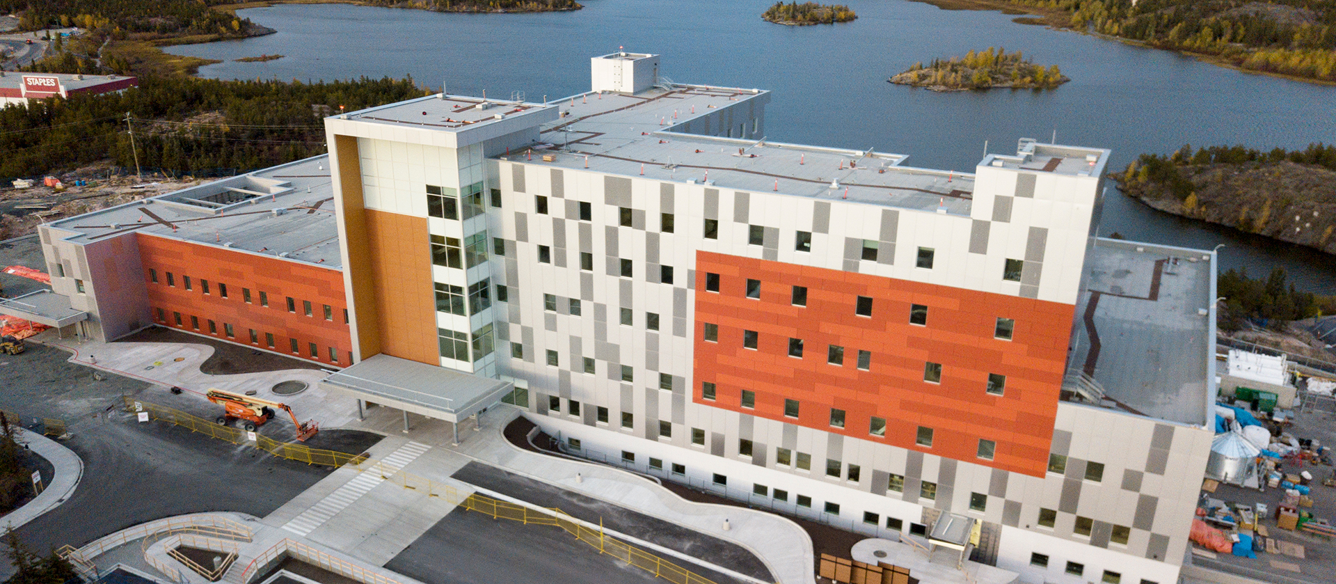 Vue aérienne d'un bâtiment hospitalier moderne de plusieurs étages, composé de panneaux blancs, gris et rouille, situé près d'un plan d'eau et entouré de parkings et de zones de construction.