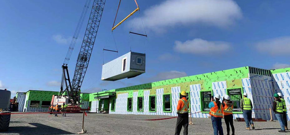 A crane lifts a prefabricated module at a construction site while workers in safety gear observe. The building under construction is partially covered in green and white insulation panels. The sky is mostly clear with a few clouds.