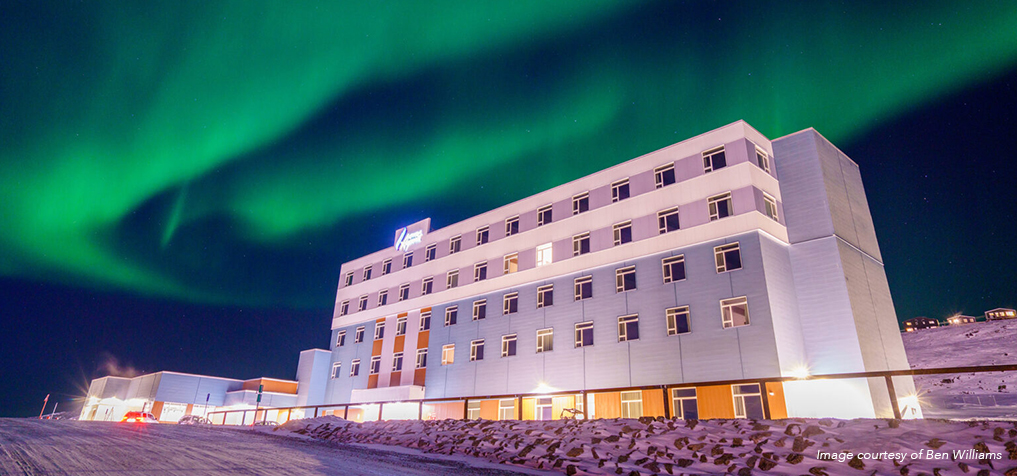 A large, modern building with many windows stands under a vivid green aurora borealis in a snowy landscape at night. The sky glows with bright northern lights above the structure.