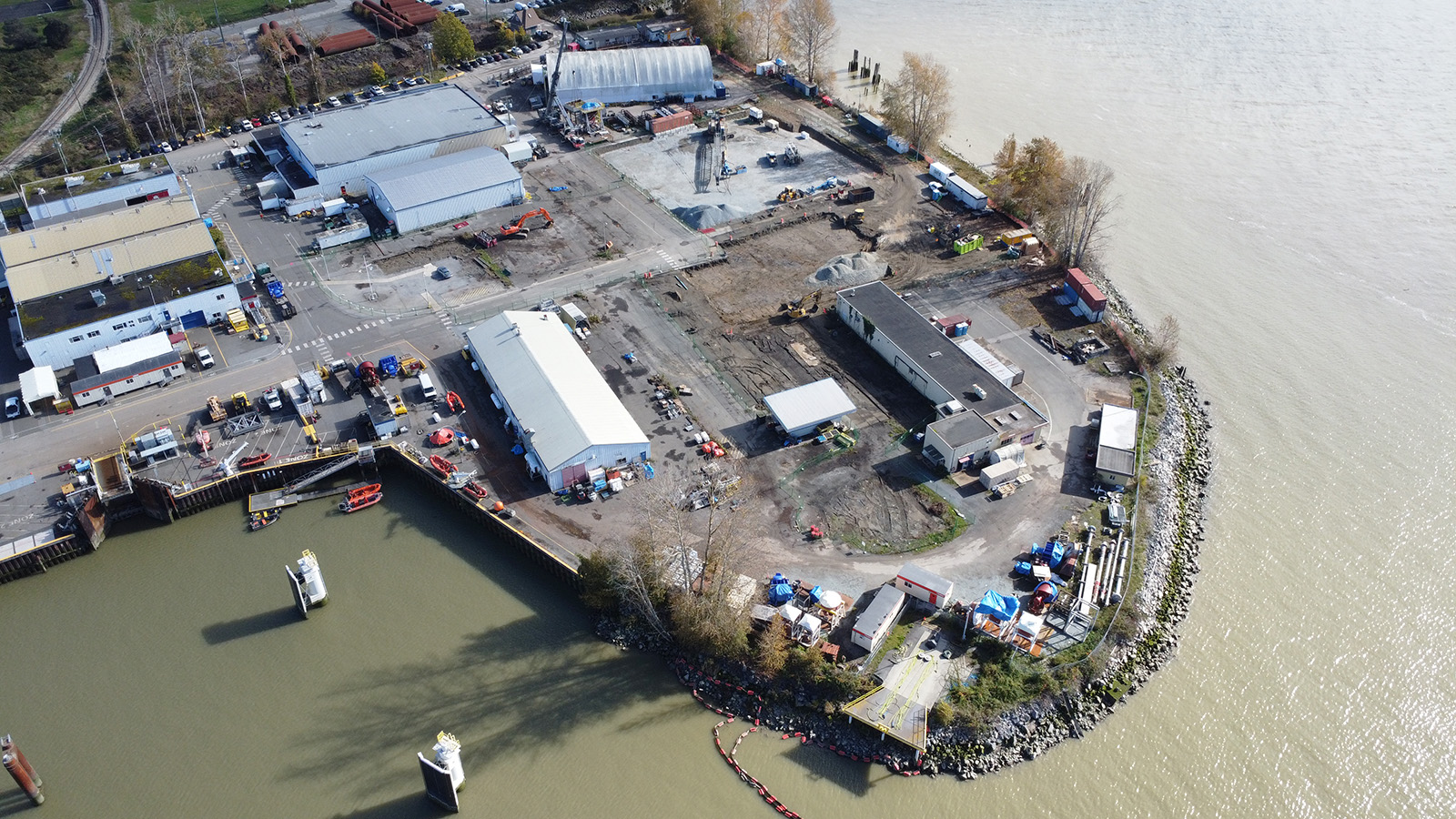 Vue aérienne d'un site industriel sur une péninsule fluviale avec plusieurs bâtiments, du matériel de construction, des véhicules en stationnement et des arbres le long de la rive. Le site est entouré d'eau sur trois côtés.