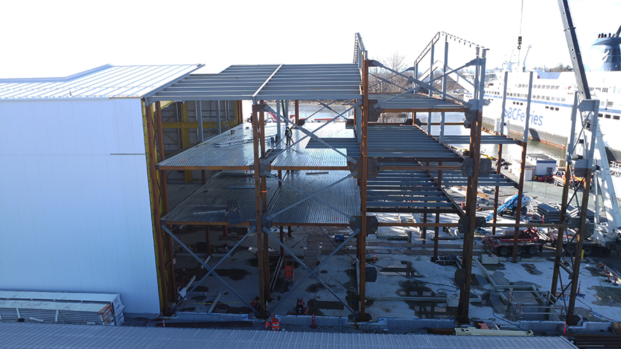 Un bâtiment à ossature métallique partiellement construit, avec plusieurs étages, des poutres ouvertes et des colonnes, est représenté sur un chantier de construction, avec des équipements et une grue visibles ; un grand ferry blanc se trouve à l'arrière-plan.