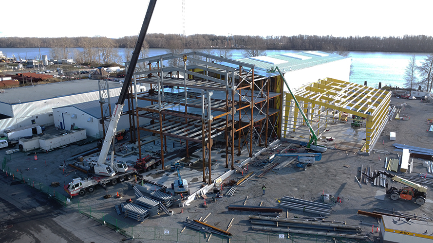 Chantier de construction avec la charpente métallique d'un grand bâtiment érigé par des grues, à côté d'une structure jaune partiellement construite, près d'une rivière et entourée d'arbres et de bâtiments industriels.