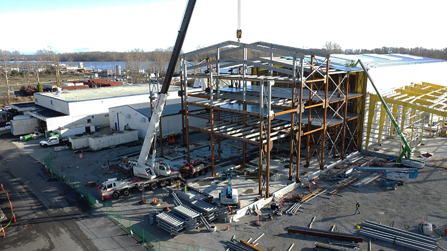 Sur un chantier de construction, la charpente métallique d'un bâtiment de plusieurs étages est assemblée à l'aide d'une grande grue ; des véhicules de construction, des matériaux et des ouvriers sont éparpillés sur le site.