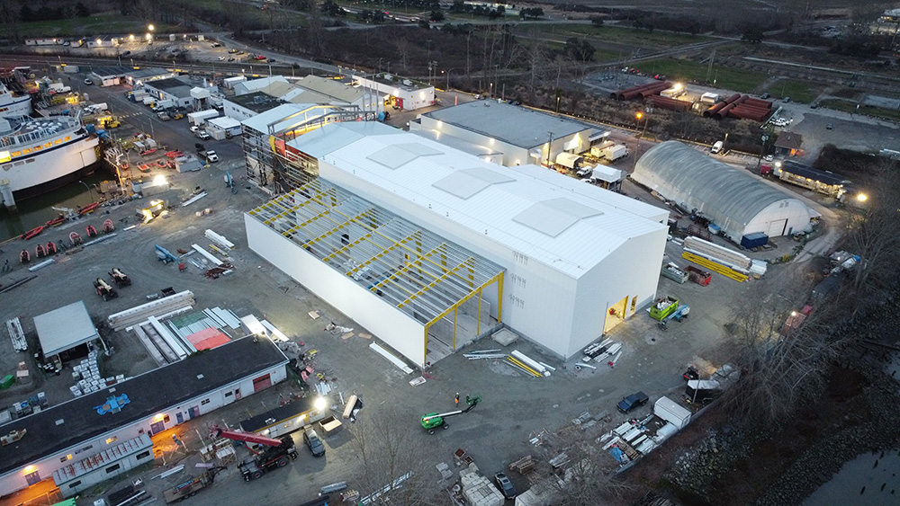 Vue aérienne d'un chantier industriel avec un grand bâtiment blanc, plusieurs véhicules, des matériaux éparpillés et des petites structures environnantes au crépuscule.