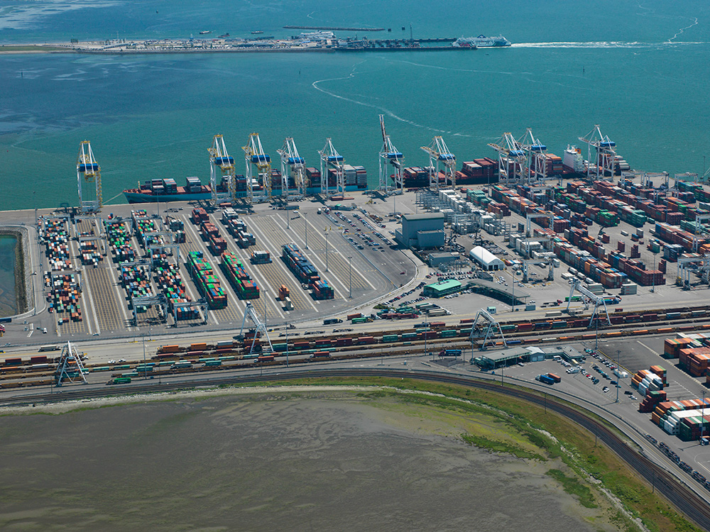 Aerial view of a busy shipping port with numerous shipping containers, cranes, and cargo ships along the waterfront, with a train track and open water in the background.