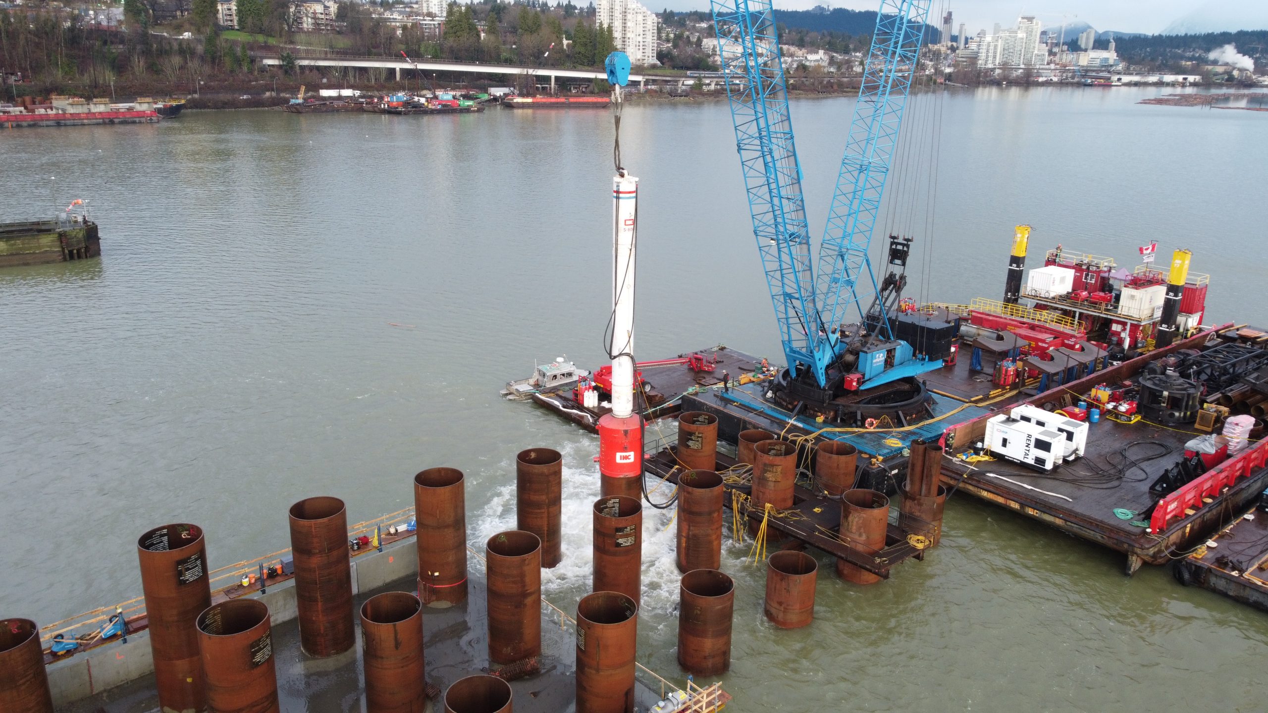 A large crane on a barge positions steel pilings into a river for construction, with multiple cylindrical pilings in the water and a cityscape with trees and buildings in the background.