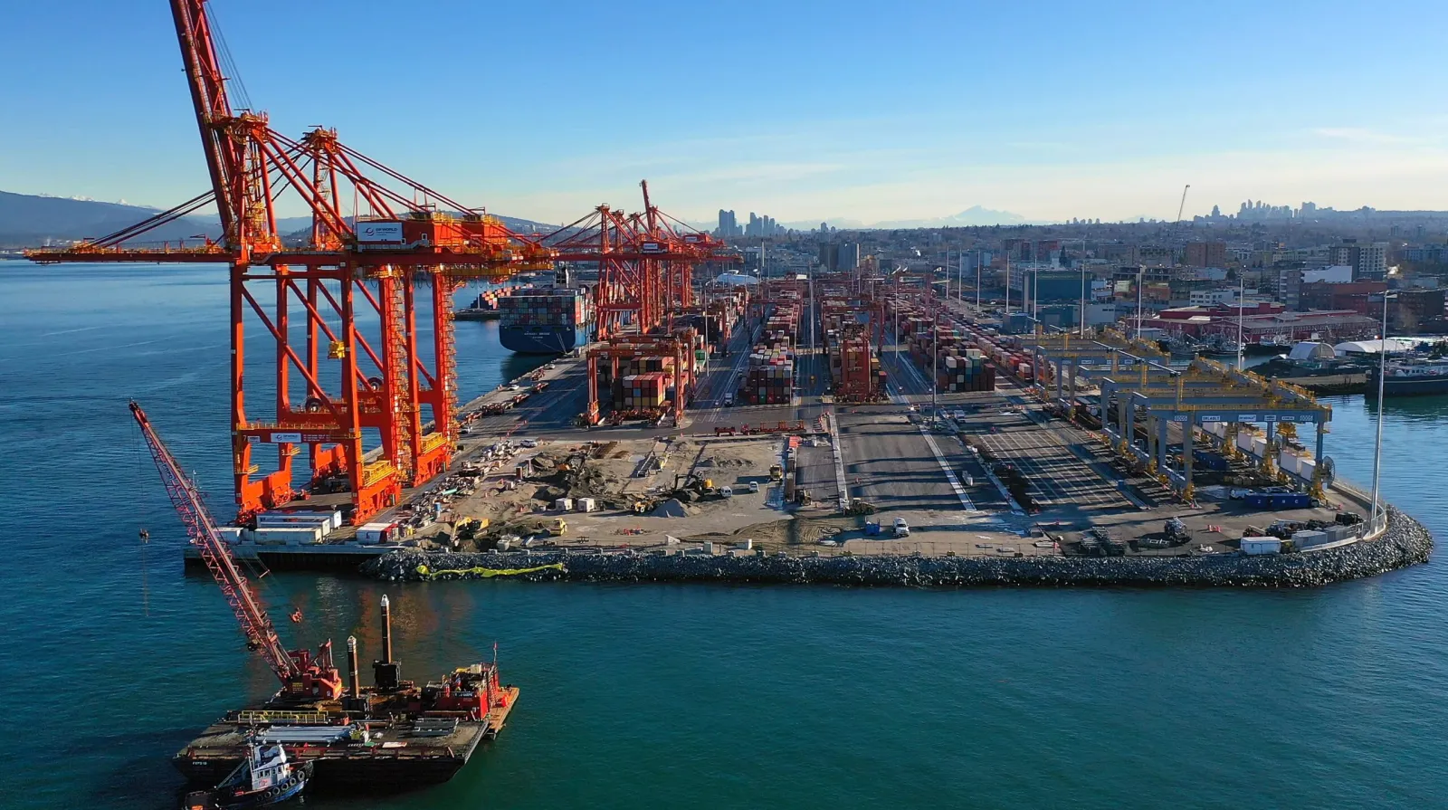 Aerial view of a busy port with large orange cranes and shipping containers along the waterfront, surrounded by water, city buildings, and a clear blue sky in the background.