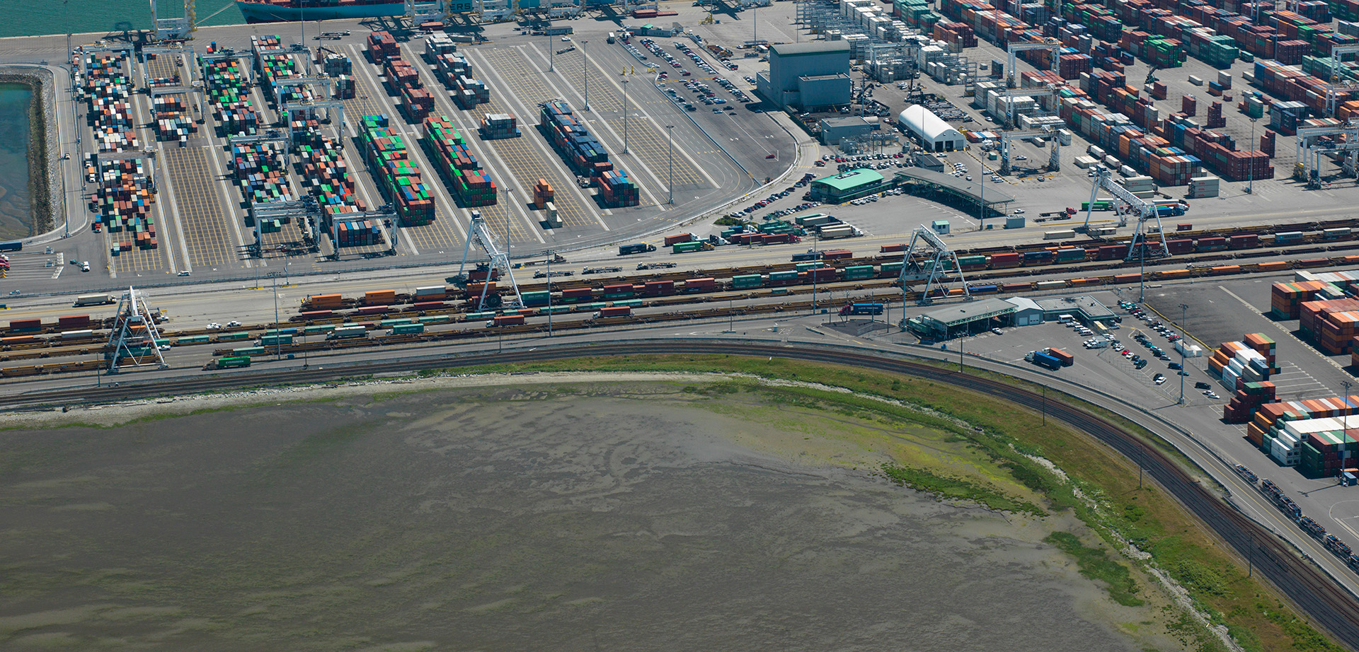 Aerial view of a busy shipping port with rows of shipping containers, cranes, trains, trucks, and storage buildings beside a body of water and a curved shoreline.