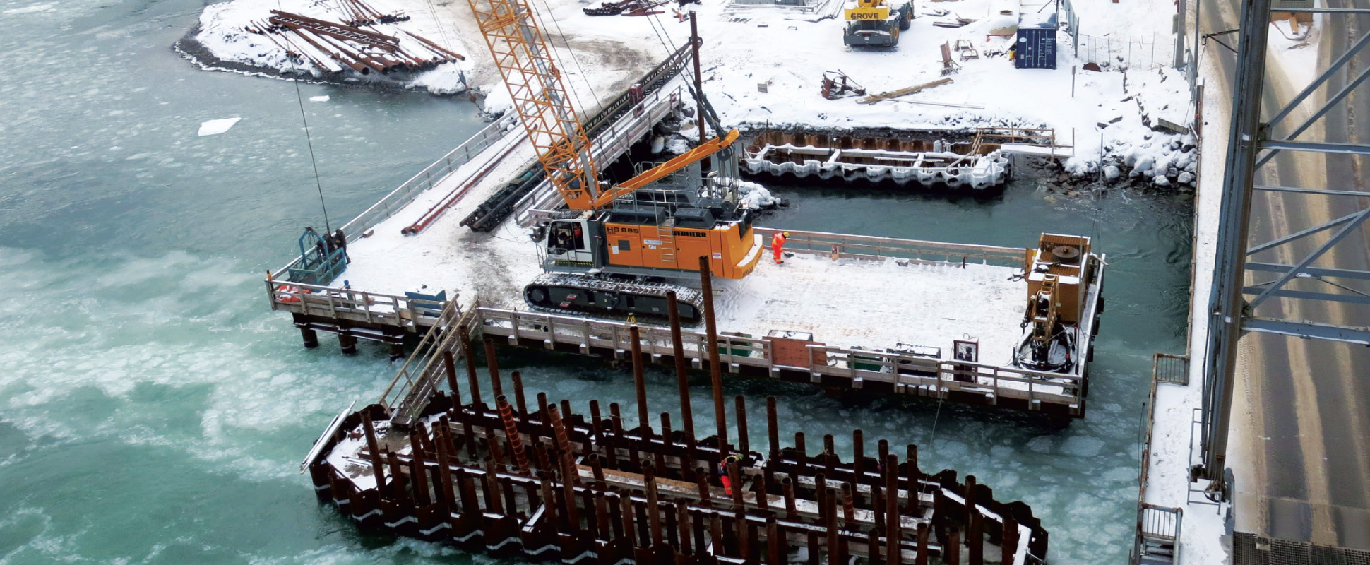 A construction site by a snowy river features a large yellow crane on a platform, surrounded by steel beams and equipment. The water is partially frozen, and snow covers the ground and structures.