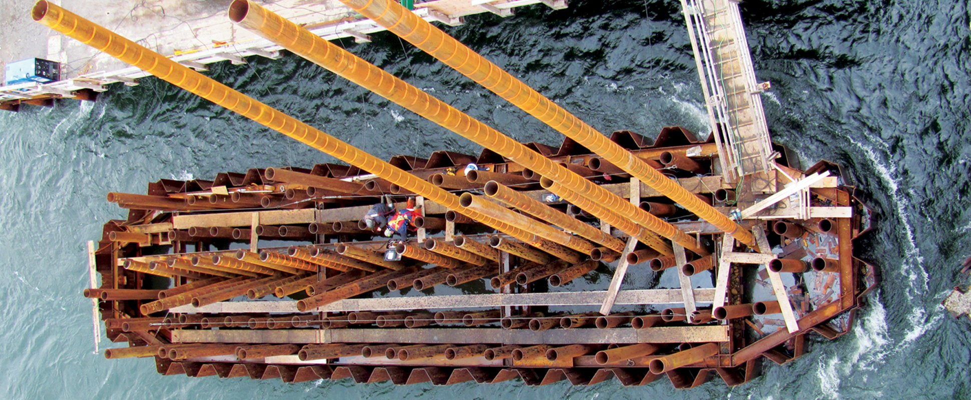 Aerial view of a construction platform in water with workers and large, vertical rusted steel beams, surrounded by scaffolding and walkways.