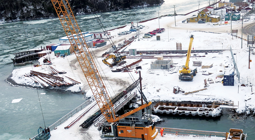 A snowy construction site by a river, featuring cranes, machinery, steel beams, and several colorful buildings in the background. The area is surrounded by water and covered in snow and ice.