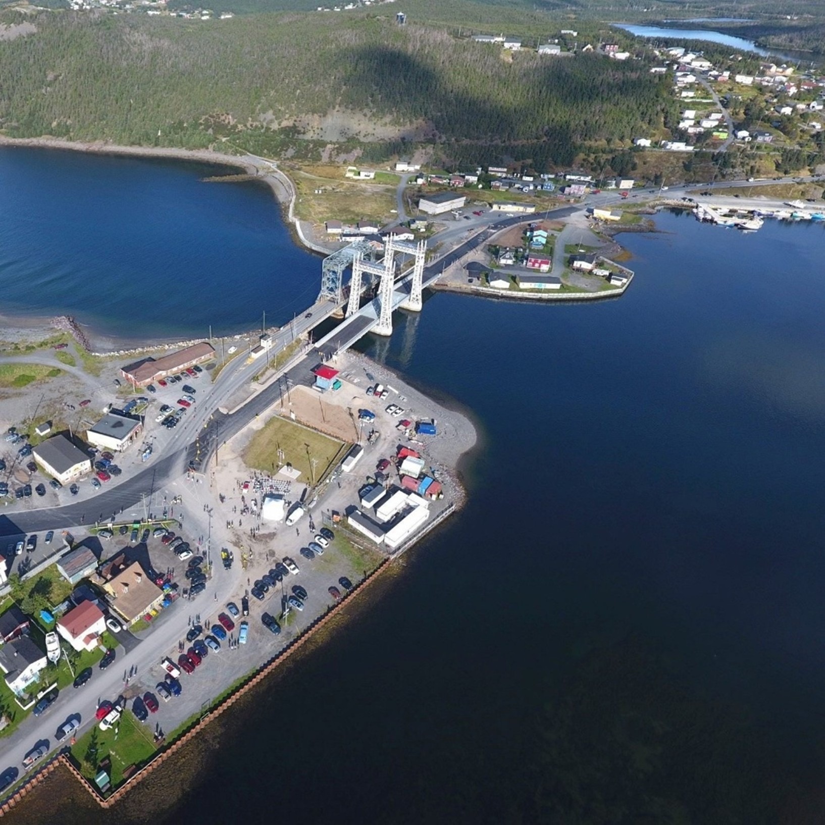 Aerial view of a small coastal town with a lift bridge spanning over water, connecting both sides. Houses, parked cars, and small buildings line the shoreline near the bridge, surrounded by green hills.