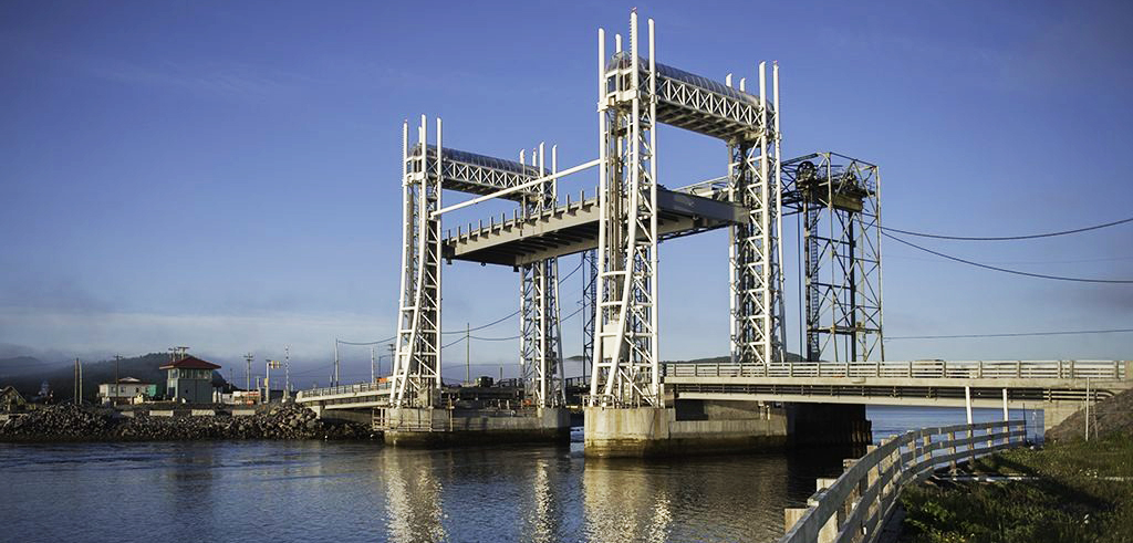 A vertical lift bridge spans over a calm river, with the central section raised. The bridge has tall white towers and is set against a clear blue sky. A small building and shoreline are visible in the background.