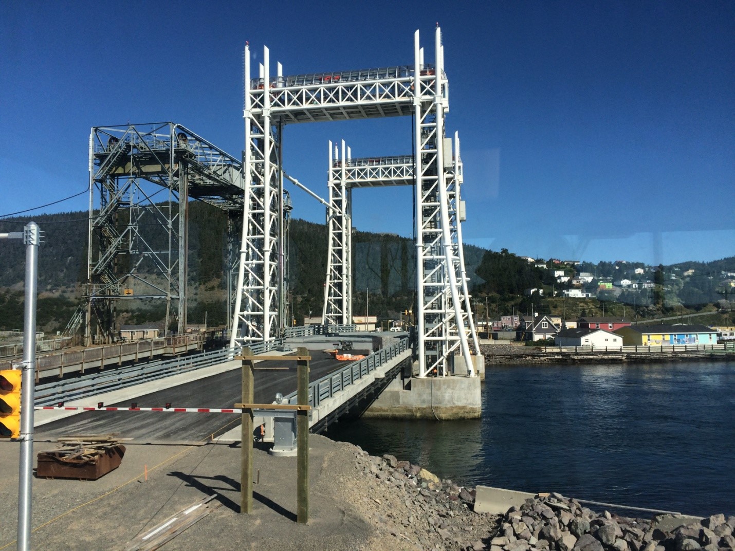 A large vertical lift bridge spans across a body of water, with its towers rising high. Houses and buildings are visible on the far shore, and the sky is clear and blue.